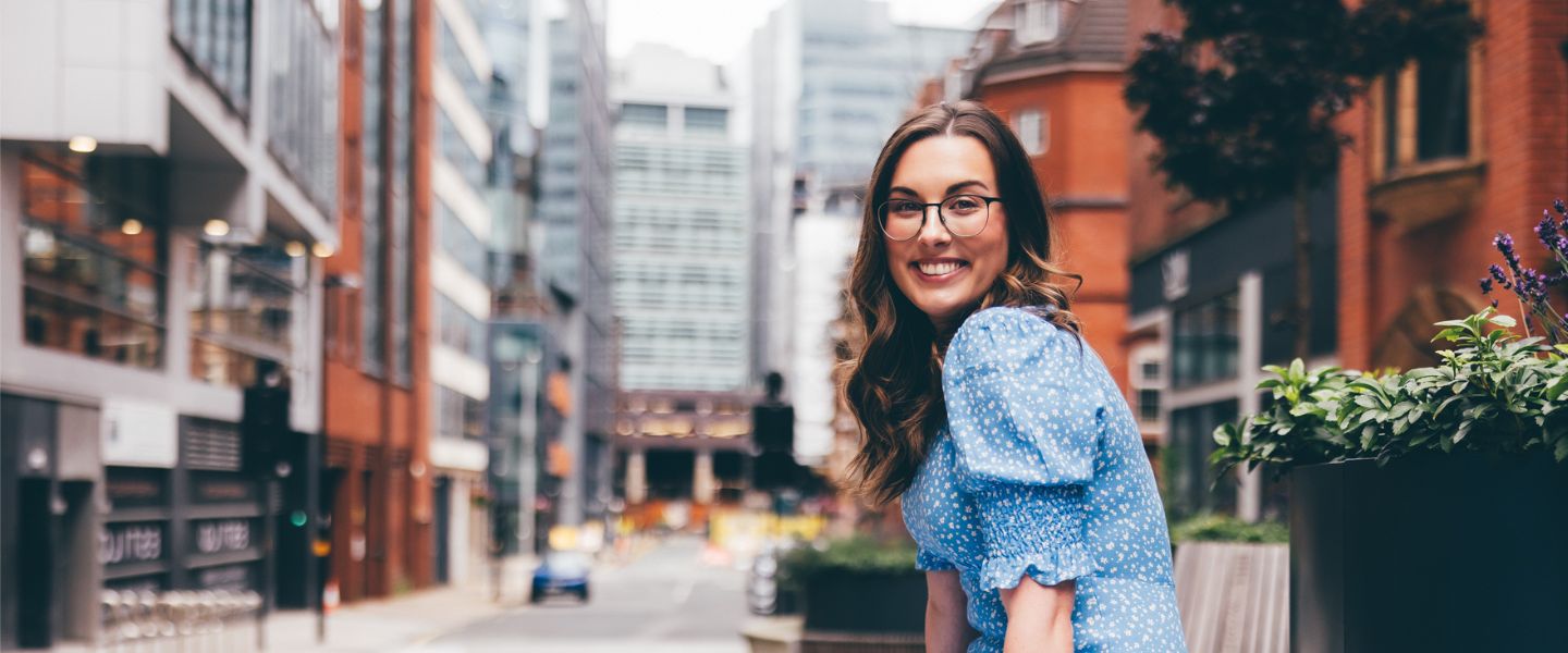 A young woman with long wavy brown hair is sat smiling at the camera, wearing a pale blue and white summer dress. She is wearing glasses and is pictured sitting outside on a bench in a city centre. 