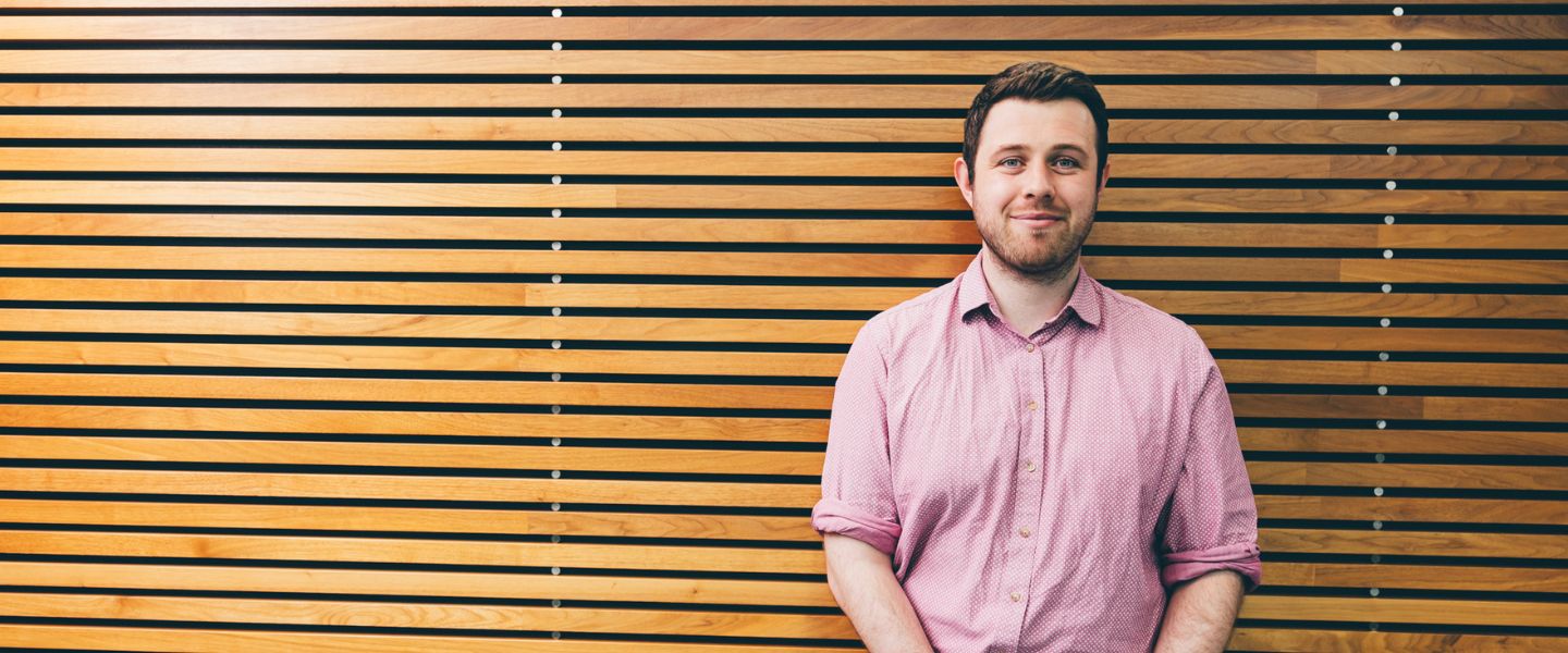 A young man in a pale pink shirt is pictured standing in front of a wooden slat wall. He has brown hair and is smiling at the camera. 