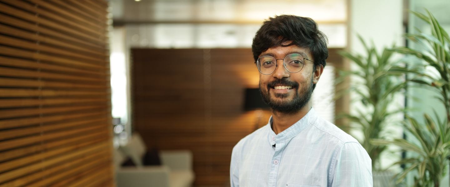 A young man is pictured from the waist up. He is standing in an open office space, with casual seating in the background. He is wearing a pale blue shirt and glasses.