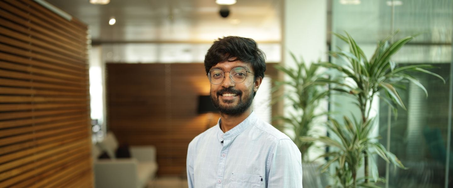 A young man is pictured from the waist up. He is standing in an open office space, with casual seating in the background. He is wearing a pale blue shirt and glasses. 