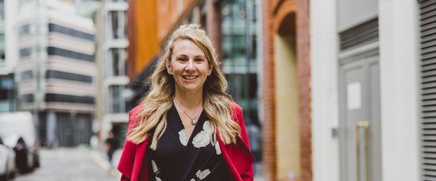 A woman with long blond hair is pictured walking towards the camera, smiling. She is wearing a black and white top with a bright red jacket.