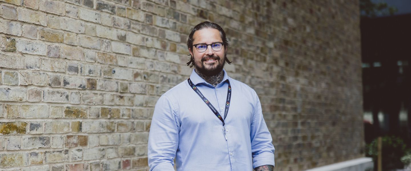 A man is smiling and looking directly at the camera. He is photographed from the waist upwards, and wearing a pale blue shirt, and a lanyard signalling neurodiversity. He has glasses and you can see tattoos at the top of his shirt, and two piercings on his chin. He is standing in front of a pale brick wall. 
