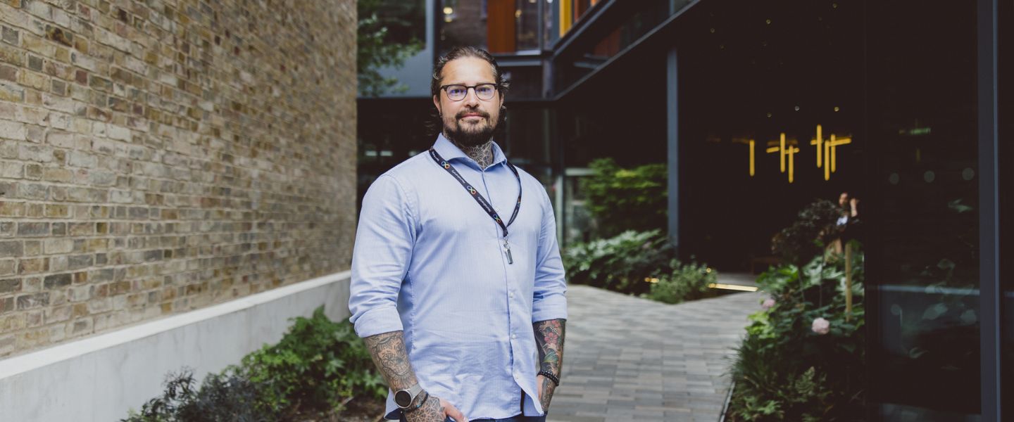 A man is smiling and looking directly at the camera, with his hands tucked into his jean pockets. He is wearing a pale blue shirt, and a lanyard signalling neurodiversity. He has glasses and you can see tattoos at the top of his shirt and on his arms, and two piercings on his chin. He is standing in front of a pale brick wall. 