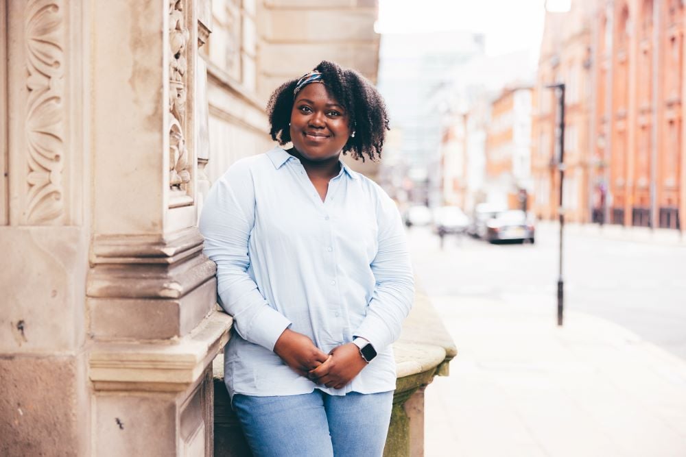 A woman is pictured in leaning against a pale brick wall, wearing a pale blue shirt and white trousers. She is smiling at the camera with a hands rested in her lap.