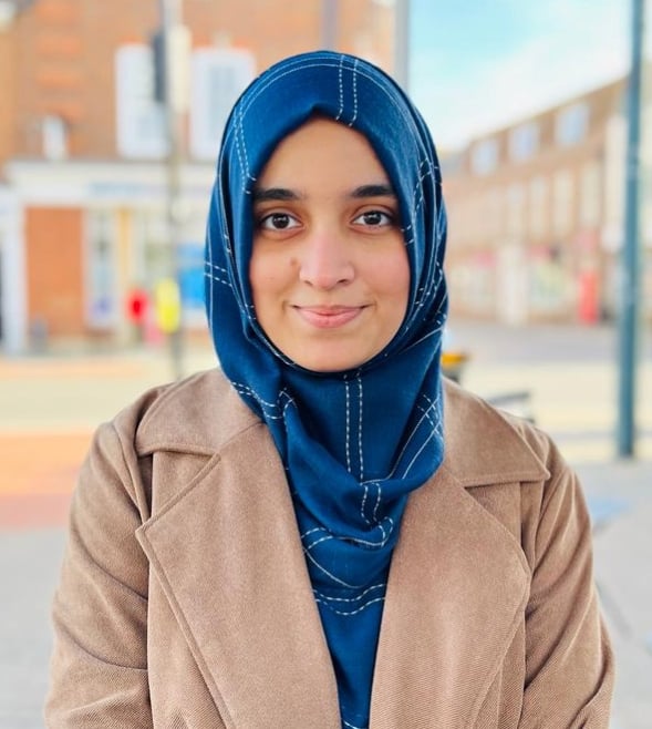 A South Asian woman wearing a blue silk hijab and beige coat is sitting outdoors. She is looking at the camera and smiling in a posed photo. She is visible from the shoulders up.