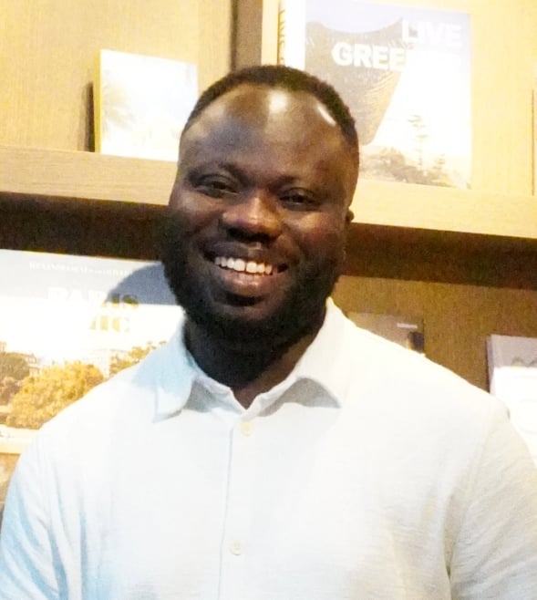 A Black man is standing in front of a bookcase inside a library, smiling at the camera. He is visible from the chest up. He has short black hair and a beard, and is wearing a white short-sleeved shirt.