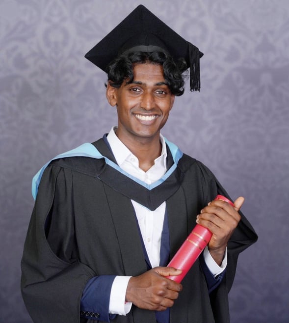A young South Asian man is posing for a formal and professional graduation photo. He is smiling at the camera, holding his degree, and wearing a cap and robes.