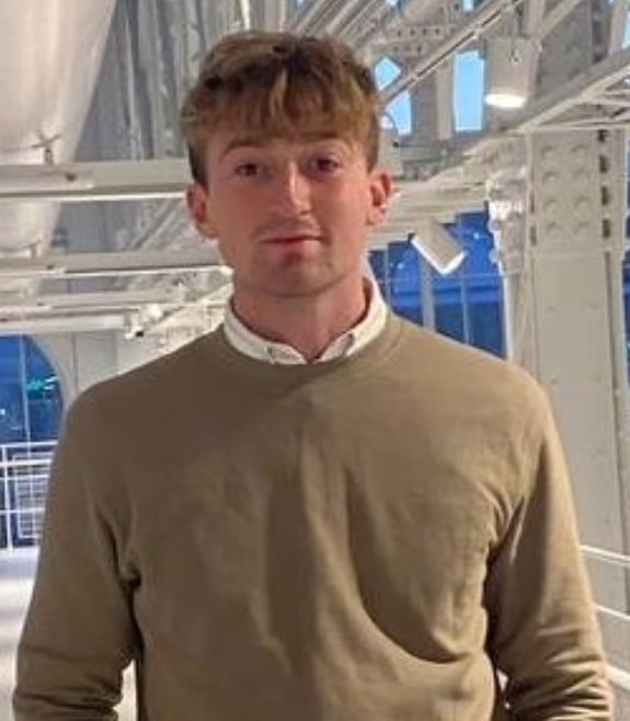 A young man with dark blonde hair is wearing a beige jumper. He's stood inside a white building that looks like a factory or manufacturer, and is looking at the camera with a slight smile. 