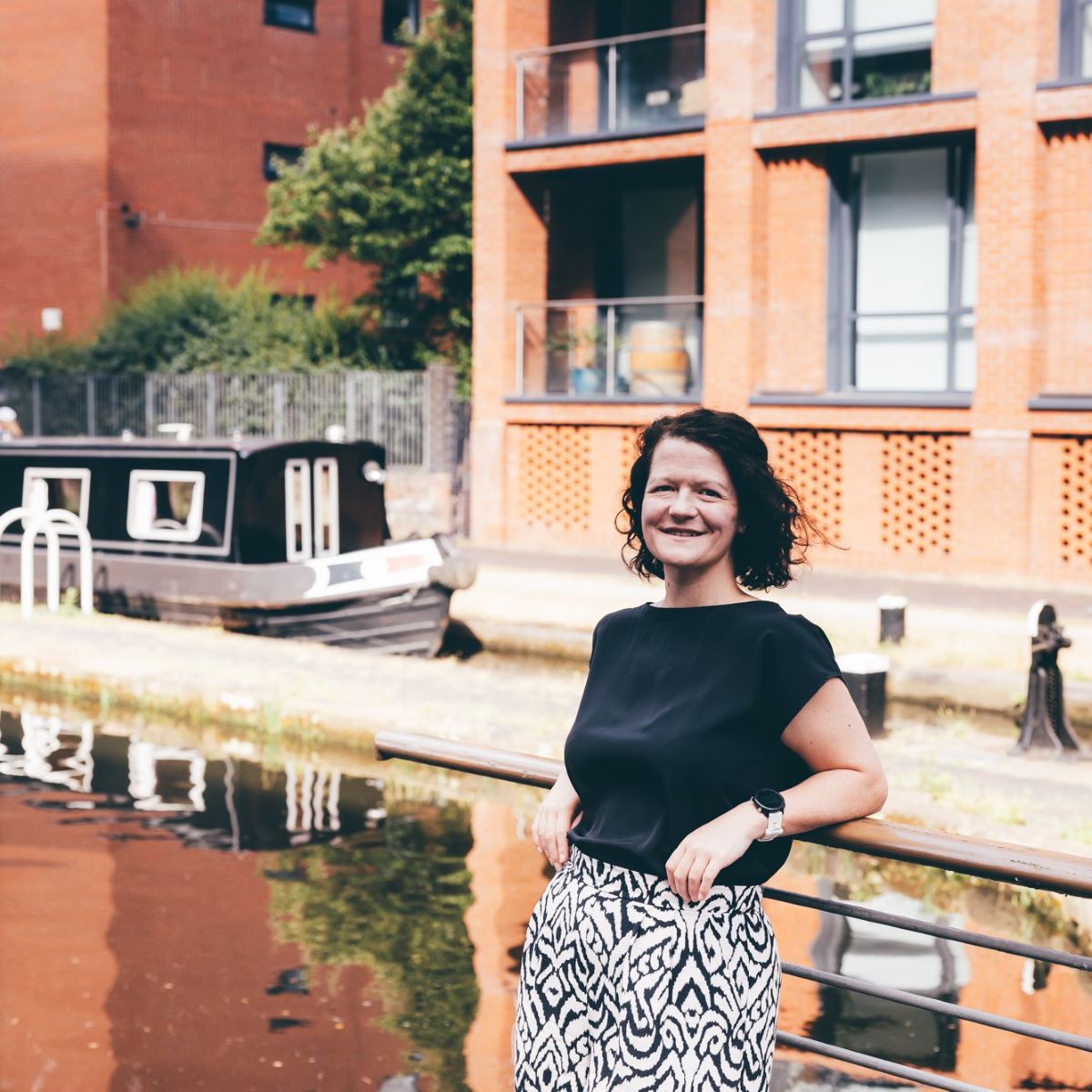 A woman with short brown hair is pictured leaning against railings by a canal. She is wearing a black short-sleeved top and black and white trousers, and is smiling at the camera. 