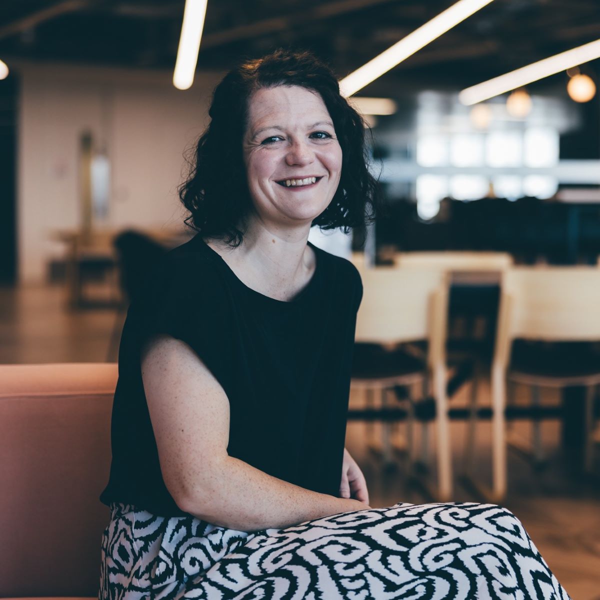 A woman with shoulder length brown hair is sat with her head turned towards the camera, smiling. She is wearing a black top, and is sat in a comfy sofa seating area within an office. 