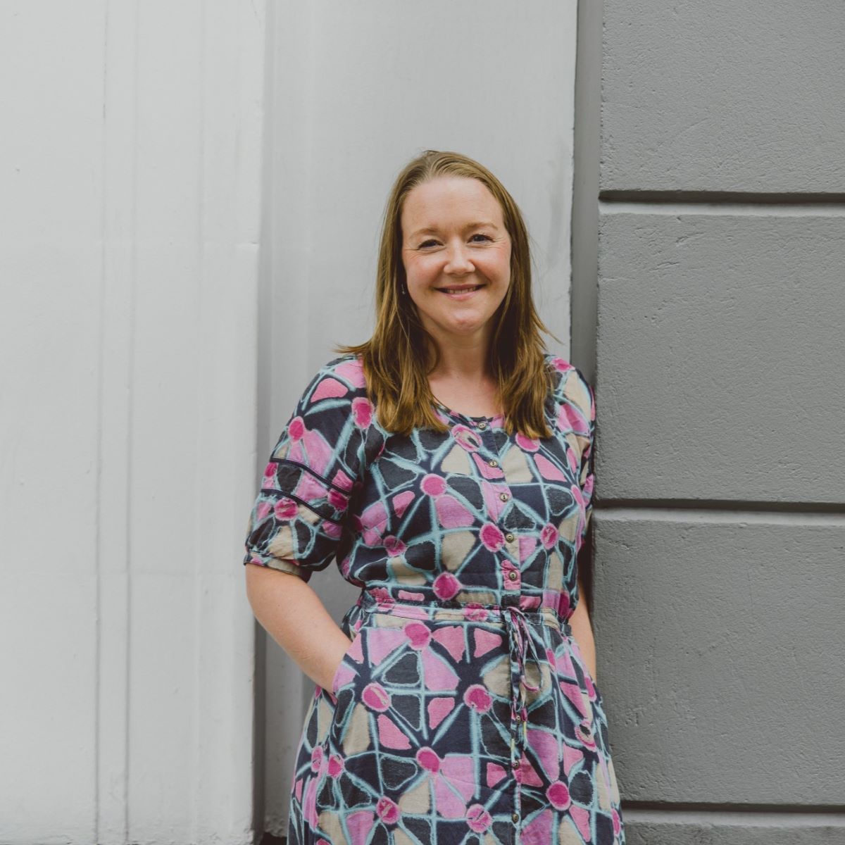 A lady is pictured standing in front of a grey brick wall. She is smiling at the camera and wearing a grey dress with brightly coloured patterns.