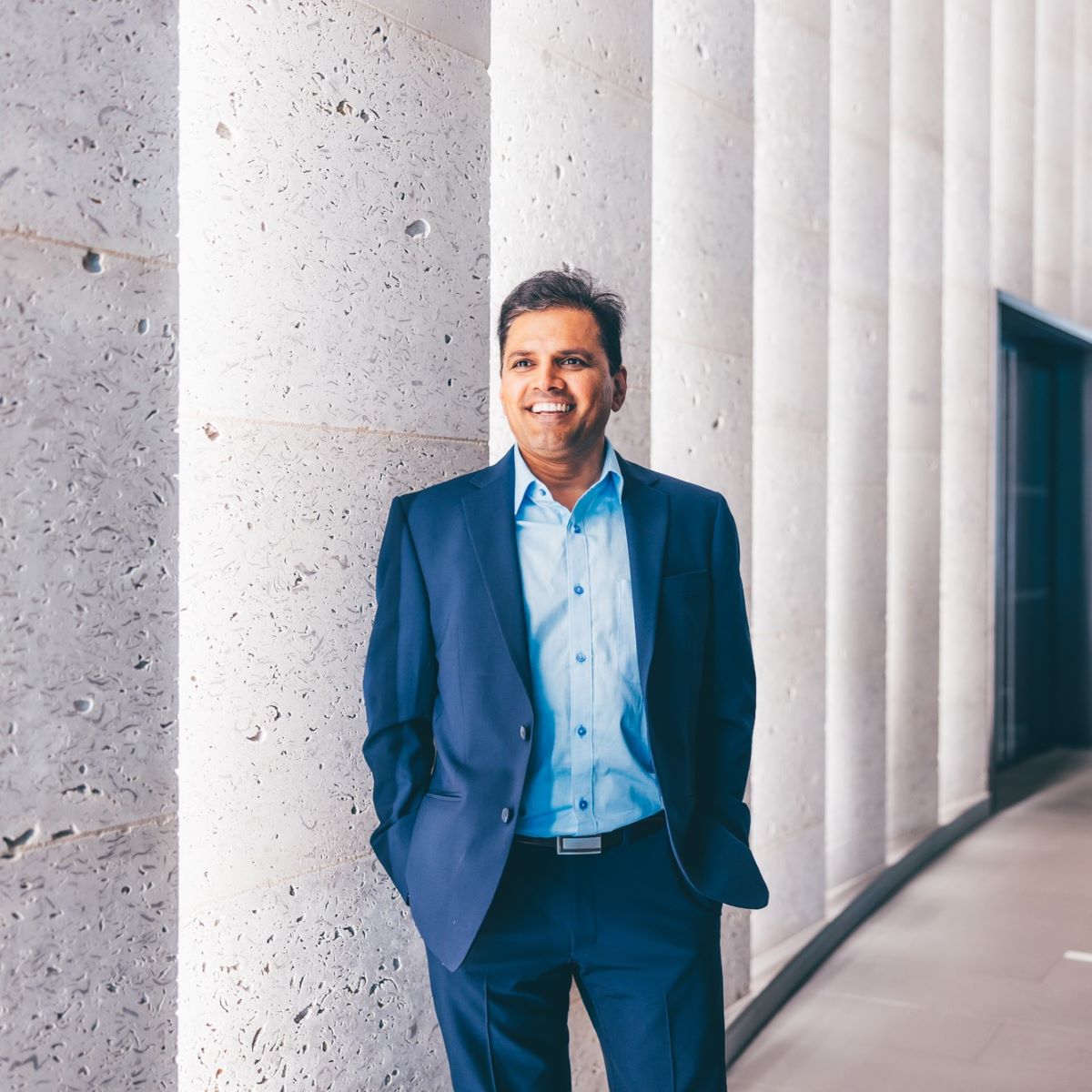 A man is pictured in an office reception, with concrete walls. He is wearing a blue shirt and a dark blue suit jacket, and is smiling at the camera.