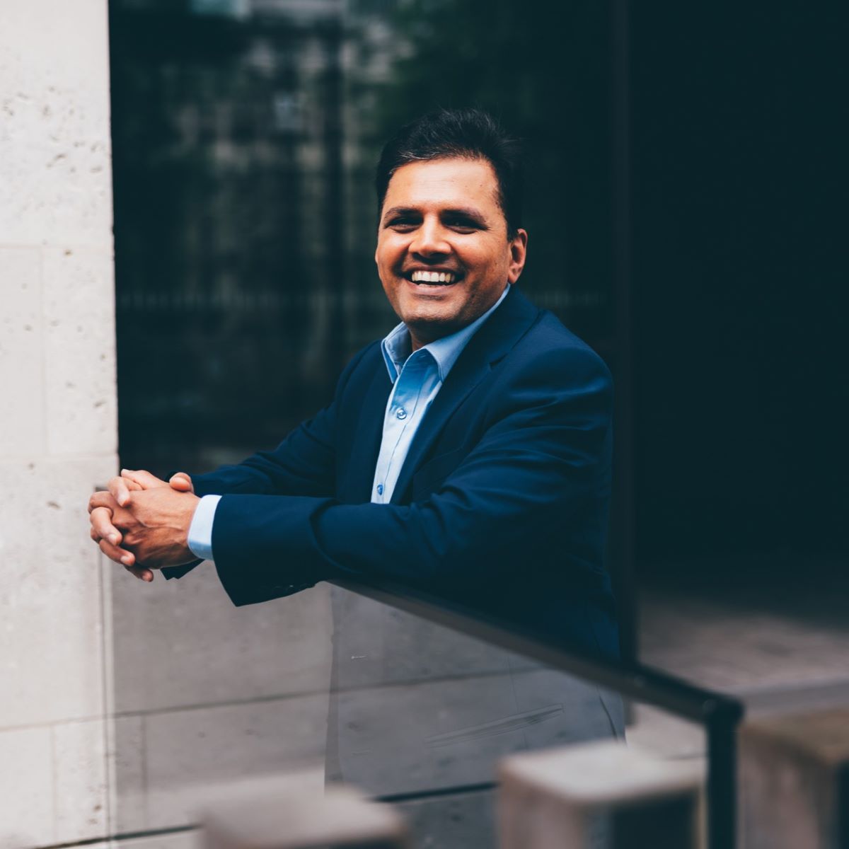 A man is pictured leaning against the railings, outside an office. He is wearing a blue shirt and a dark blue suit jacket, and is smiling at the camera. 
