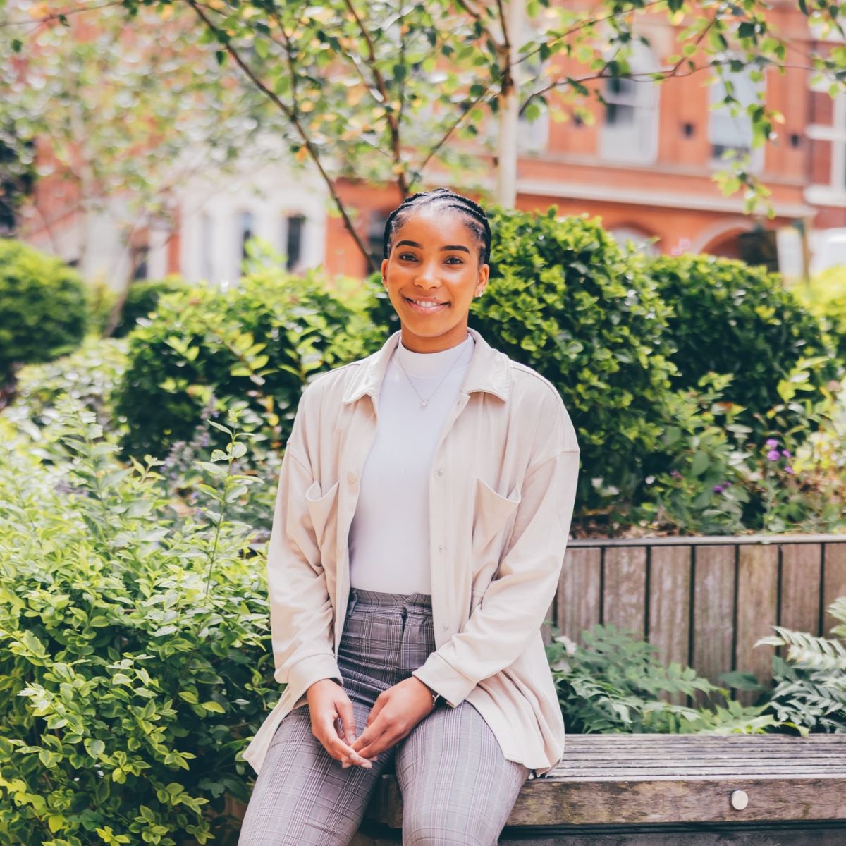 A young woman is pictured leaning against a raised flower bed in a city space with dark-brick buildings in the background. She is looking directly at the camera and smiling, wearing grey tailored trousers and a pale cream top and shirt.