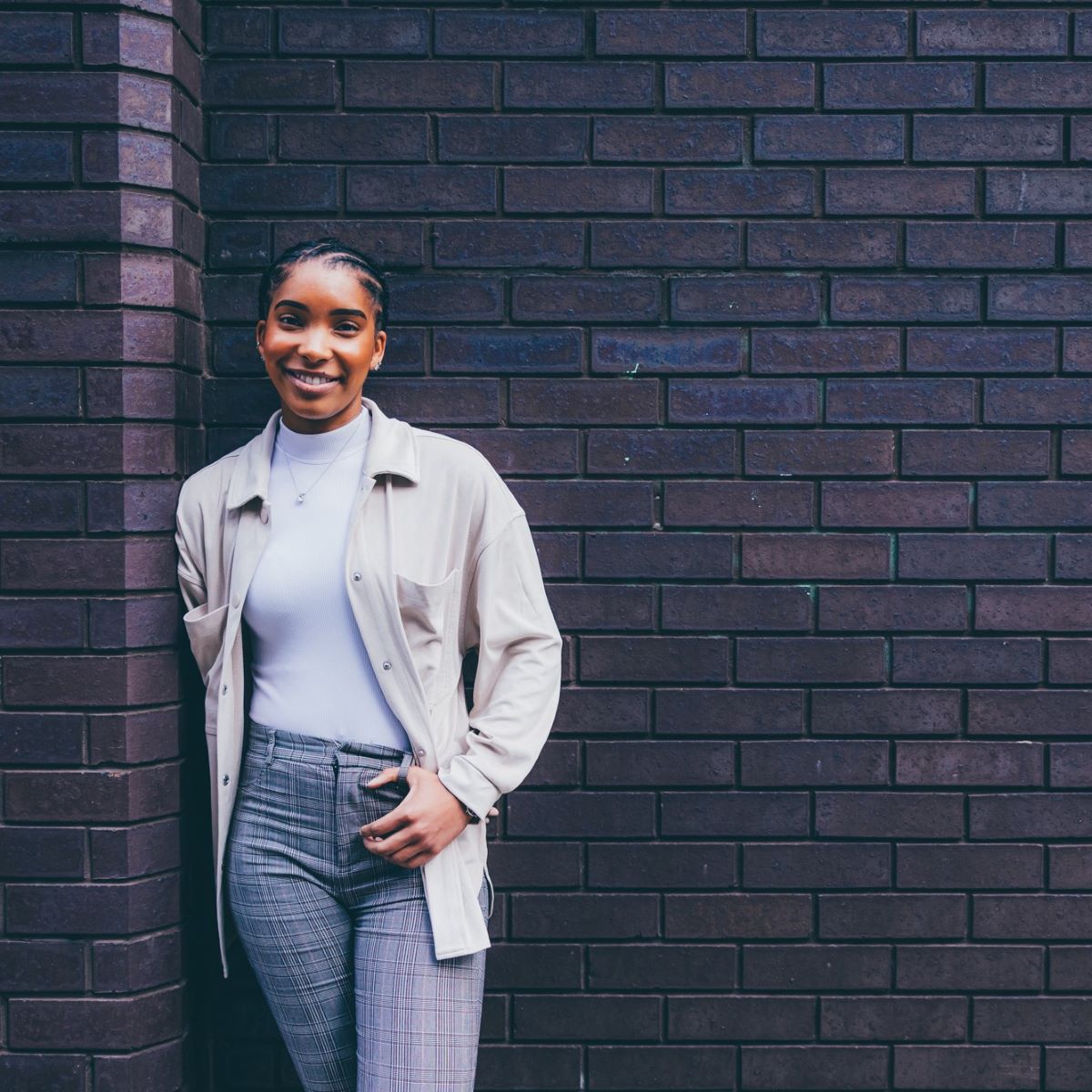 A young woman is pictured leaning against a dark brick wall. She is looking directly at the camera and smiling, wearing grey tailored trousers and a pale cream top and shirt.