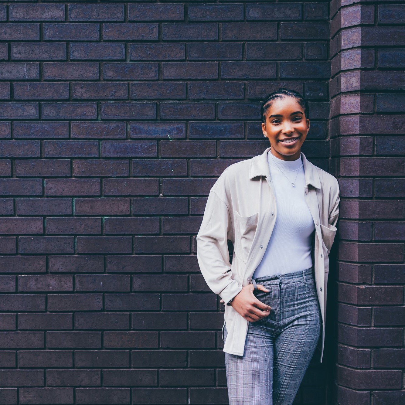 A young woman is pictured leaning against a dark brick wall. She is looking directly at the camera and smiling, wearing grey tailored trousers and a pale cream top and shirt.