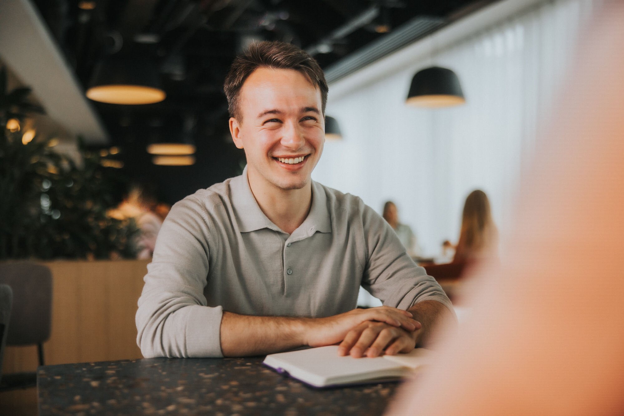 A young man with short blonde hair and a beige polo shirt is sat at a cafeteria table. He is looking at someone in front of him who isn't seen on camera. He is smiling.