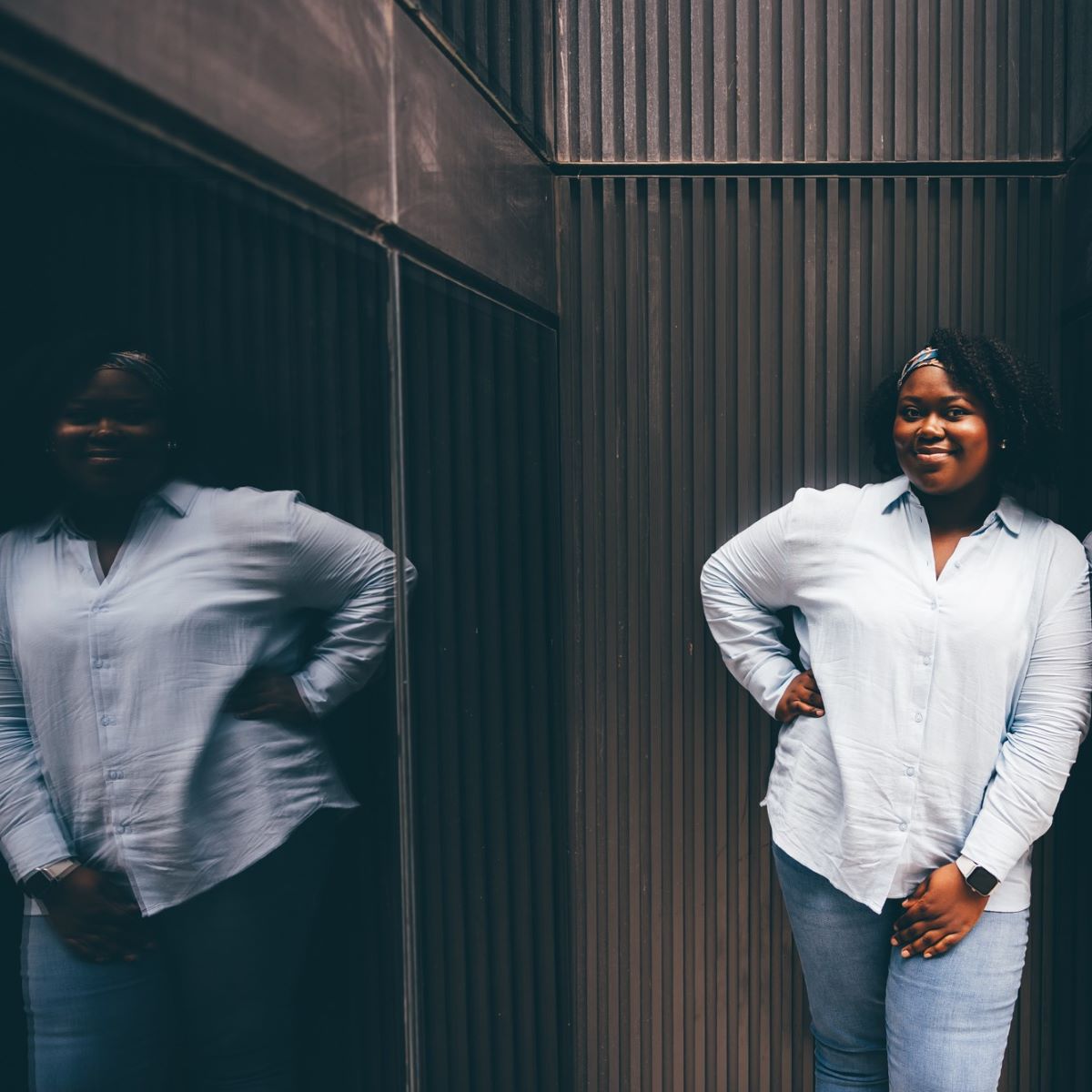 A woman wearing a pale blue shirt, smiling at the camera with a hand on her hip. She is photographed outside, against a black brick wall and black glass wall. The black glass wall gives a mirrored reflection of the woman. 
