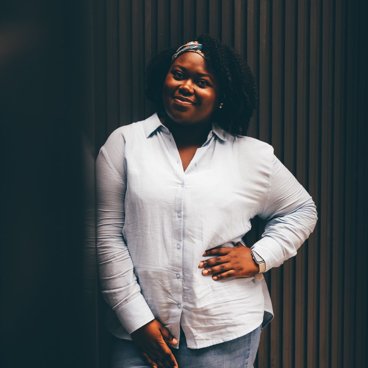 A woman wearing a pale blue shirt, smiling at the camera with a hand on her hip. She is photographed outside, against a black brick wall and black glass wall. 