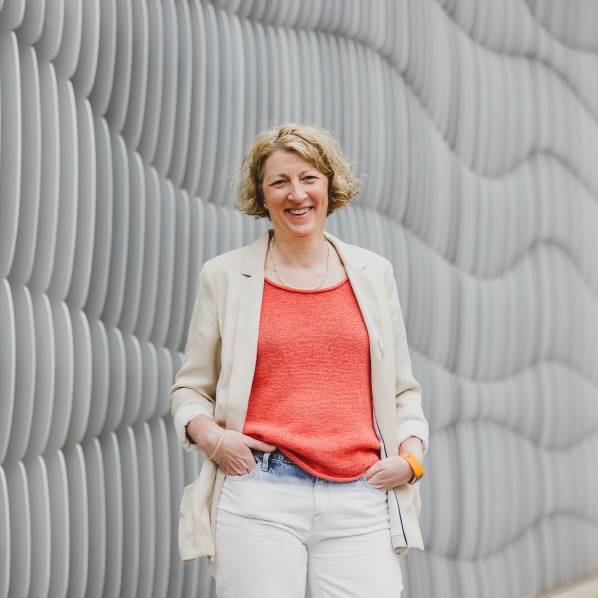 A woman with short cropped hair pictured standing outside wearing a coral top and beige jacket.  She is smiling at the camera, in front a silver building.