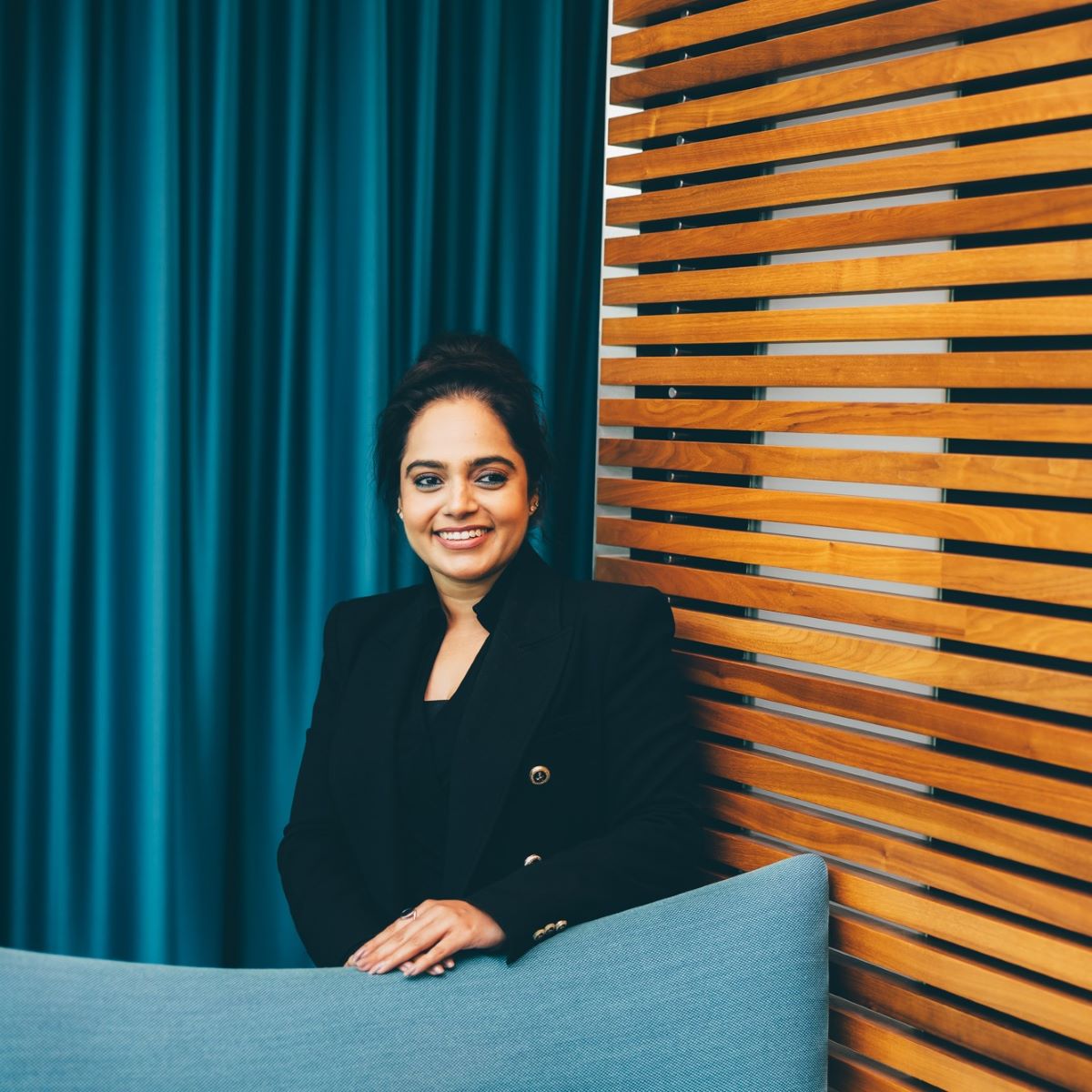 A woman is pictured standing behind a high-backed teal office chair. She is wearing a black suit and glasses and is smiling whilst looking off camera. 