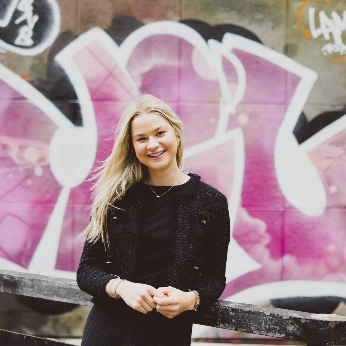 A young woman with long blond hair is photographed wearing a black top, stood against a fence in front of bright pink graffiti. 