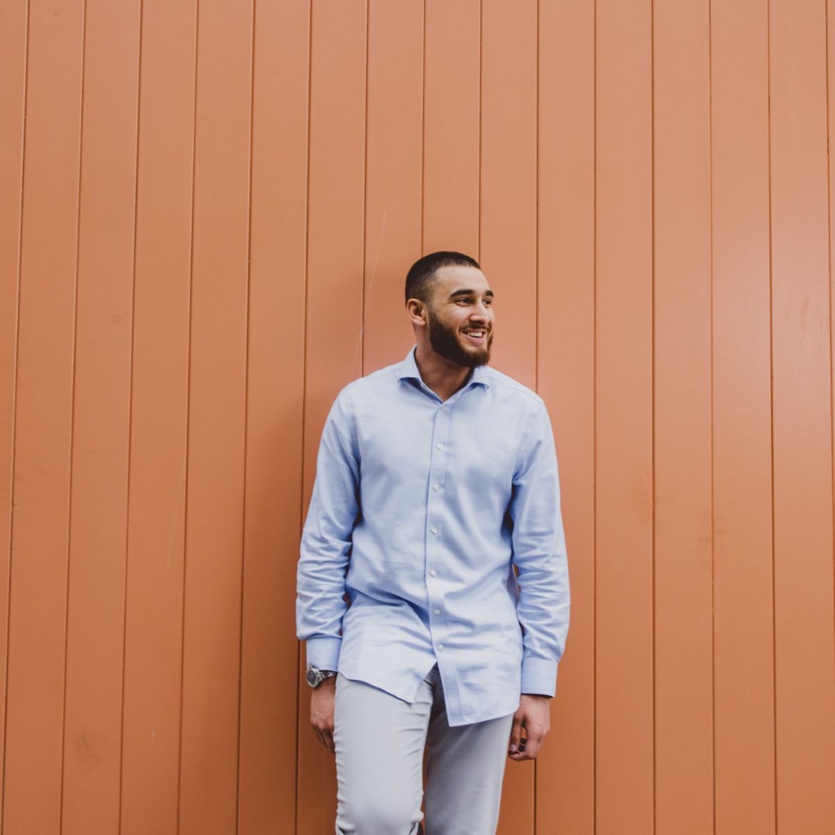 A young man is stood leaning against a peach coloured wooden high wooden door. He is wearing a pale blue shirt, and smiling whilst looking off camera to the right.