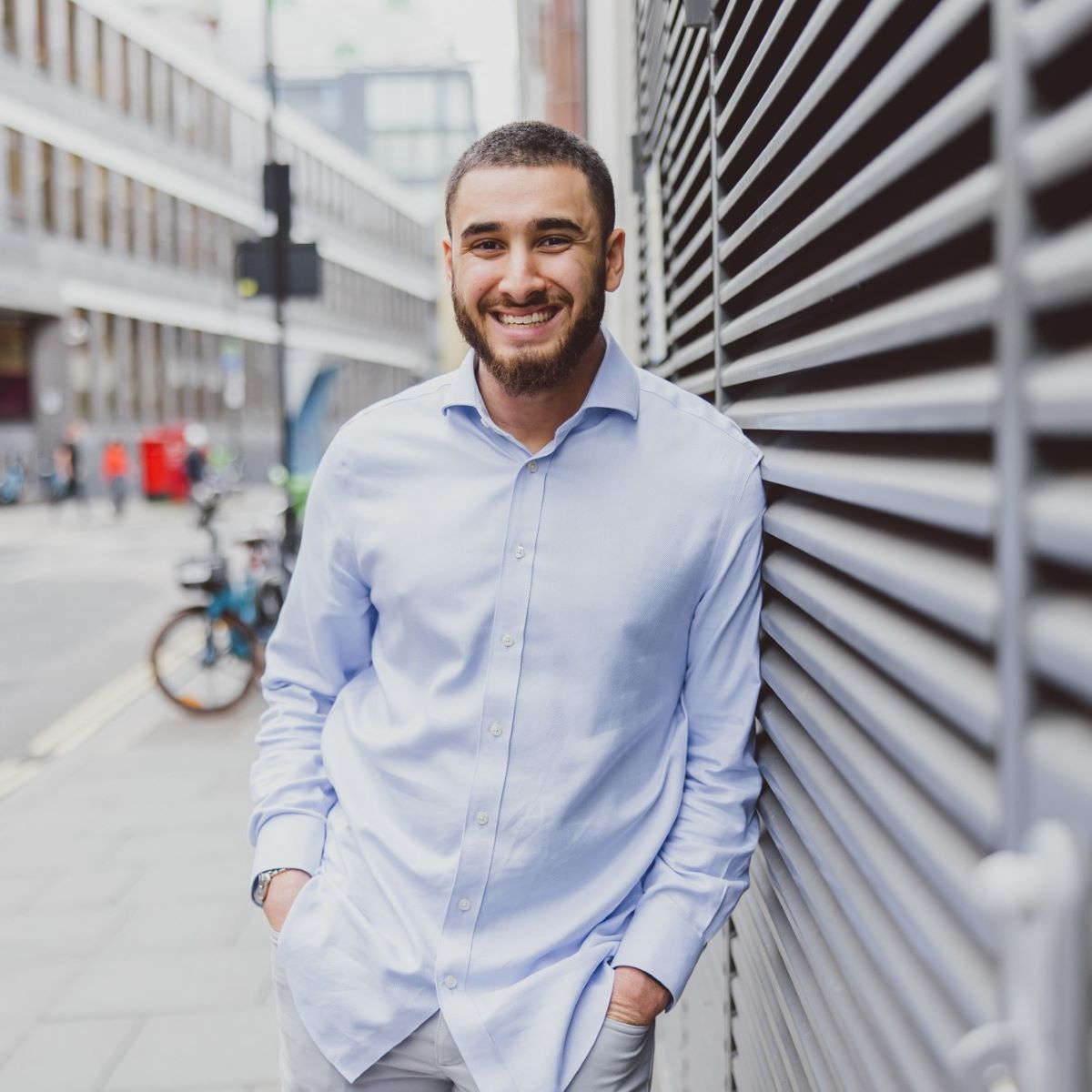 A young man is stood leaning against a building, with a busy street behind him. He is wearing a pale blue shirt, and smiling whilst looking at the camera.