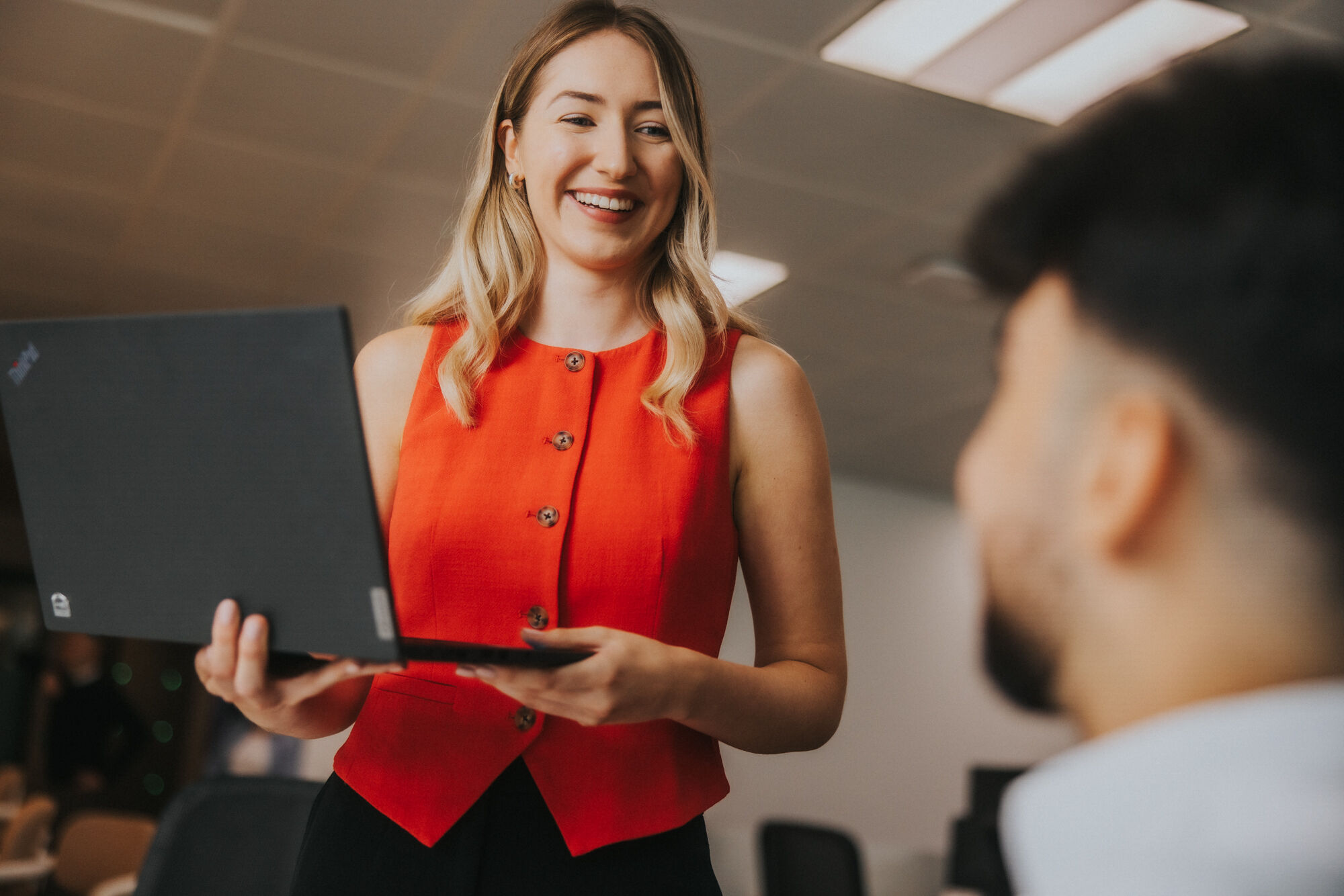 A young woman is holding a laptop and standing in front of a young man who is off camera and sat down, in an office environment. She is smiling, has long blonde hair and is wearing a red buttoned-up vest top.