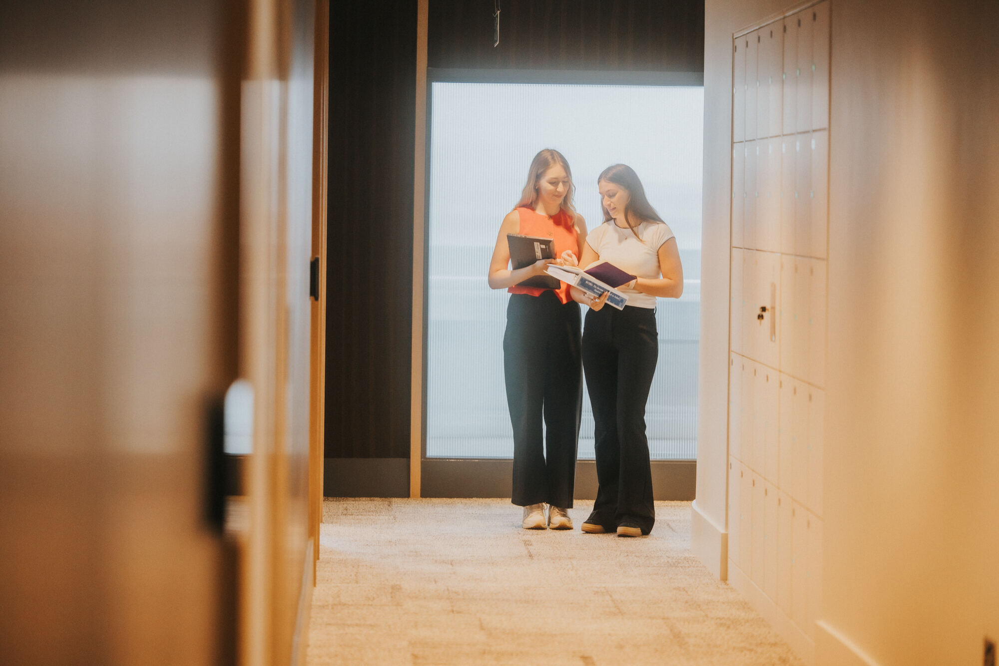 Two women stand at the beginning of a long corridor. One is blonde wearing a red top and black trousers, and the other is brunette with a white t-shirt and black trousers. They're reviewing some papers together.
