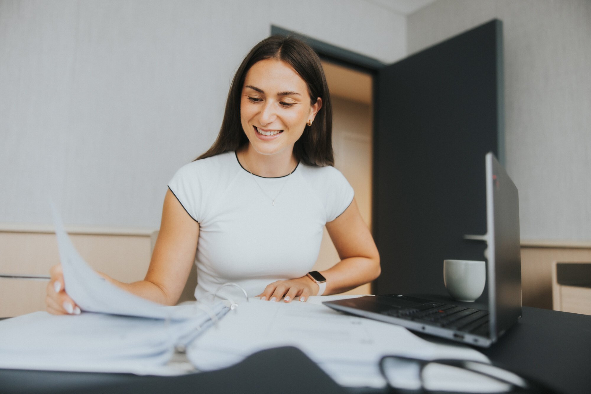 A woman with long brown hair sits at a desk looking through papers, smiling.