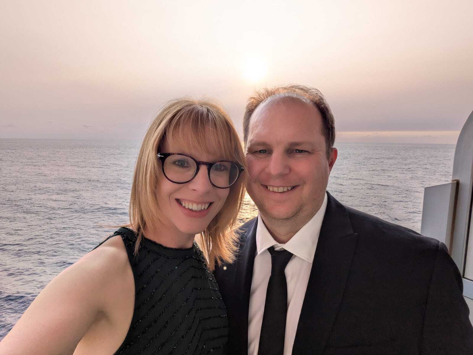 A middle aged man and woman stand in front of a sunset, smiling at the camera. The man is wearing a formal black suit and tie and the woman is wearing glasses and a black formal dress.