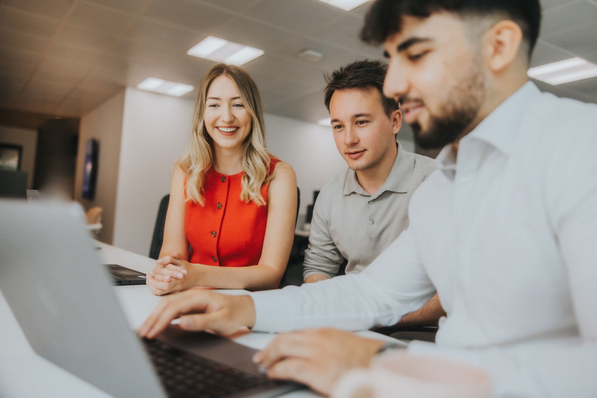 Three trainees are sat in an office building at a desk, looking at the same laptop screen. One trainee is a woman with long blonde hair and a red formal vest. One male trainee has short brown hair and a beige polo shirt. The second male trainee has a white shirt and dark brown short hair and a beard.