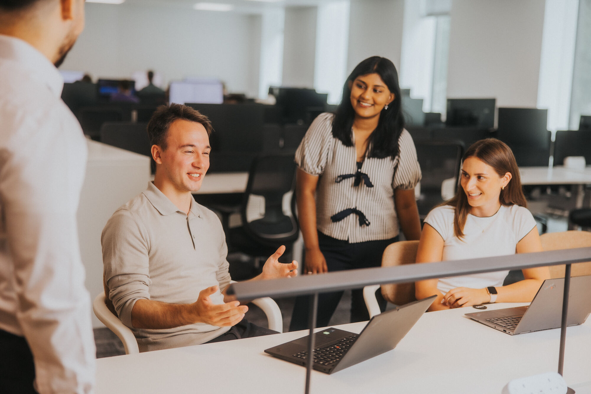 Four trainees, two male and two female, are both stood and sat around a desk in an office, smiling and appearing to talk to each other.