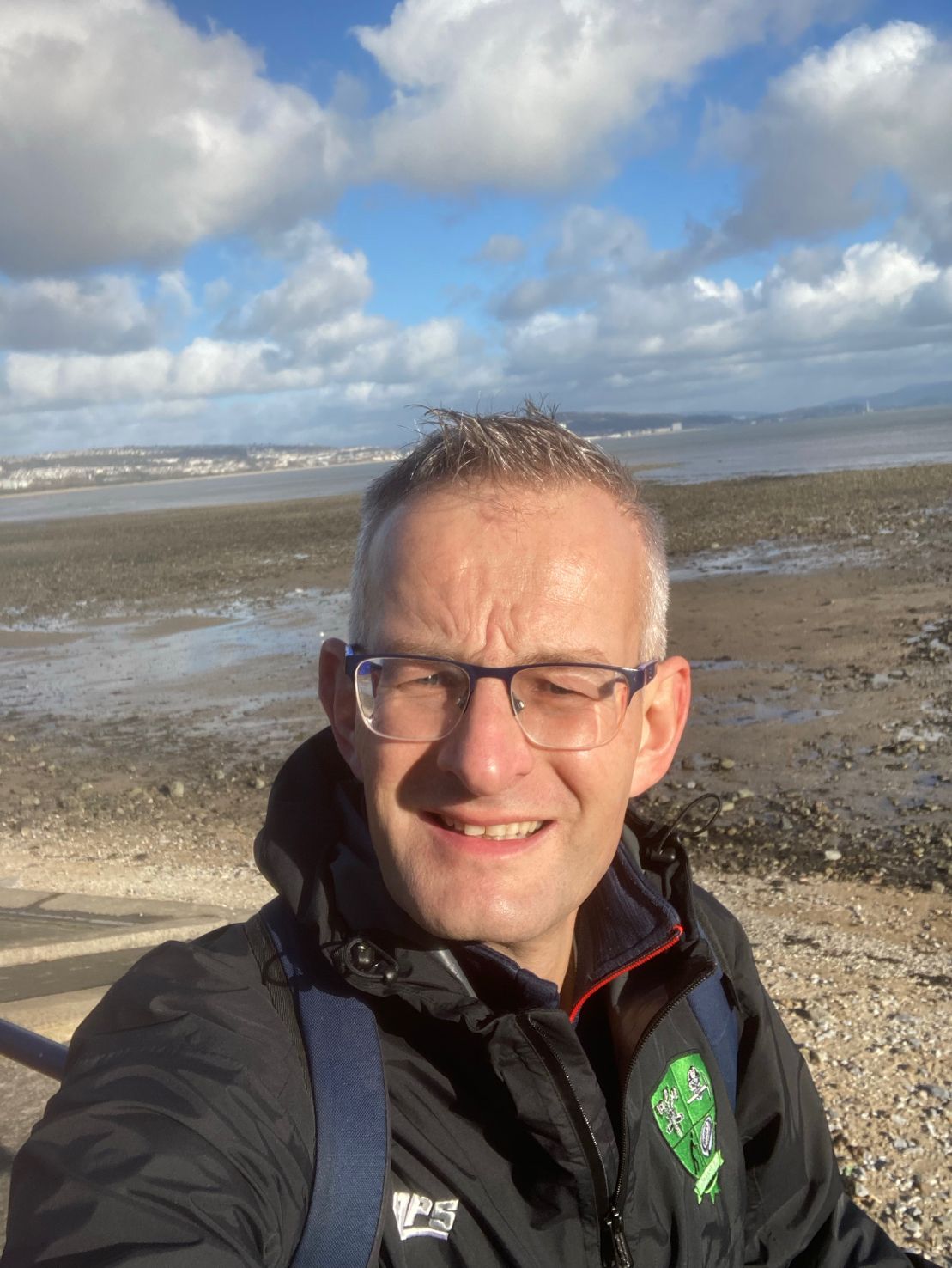 A man wearing glasses, a coat and a scarf stands outside on the beach with blue sky behind him. 